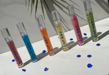 Load image into Gallery viewer, Glitter gloss collection, fuchsia, bright blue, orange yellow, pink and green lip gloss, leaves back ground and blue gems on the floor